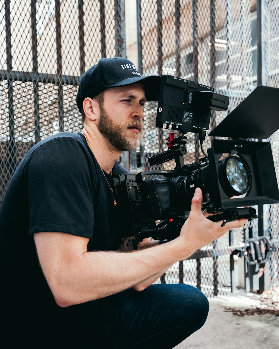 Filmmaker crouches outdoors operating a professional video camera.